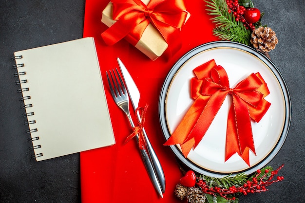 Top view of New year background with red ribbon on dinner plate cutlery set decoration accessories fir branches next to a gift and spiral notebook on a red napkin