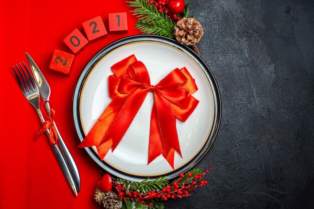 Вид сверху новогоднего фона с красной лентой на обеденной тарелке, набор столовых приборов, аксессуары для украшения еловых веток и цифр на красной салфетке на черном столе