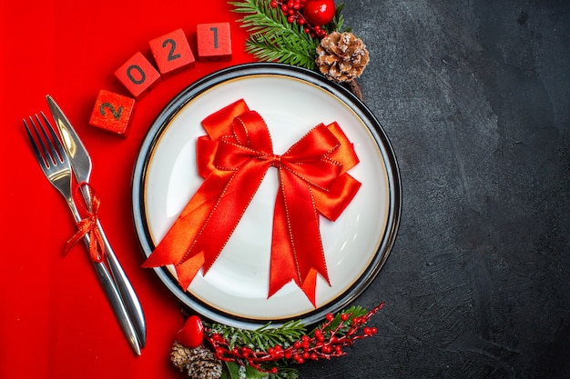 Top view of New year background with red ribbon on dinner plate cutlery set decoration accessories fir branches and numbers on a red napkin on a black table