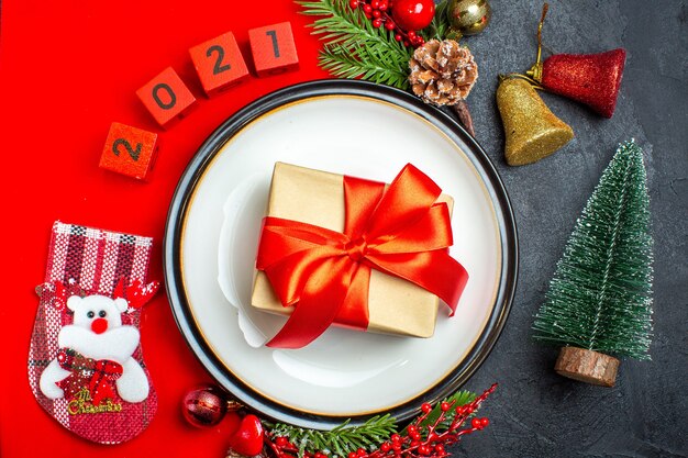 Top view of New year background with red ribbon on dinner plate decoration accessories fir branches and numbers christmas sock on a red napkin next to christmas tree on a black table