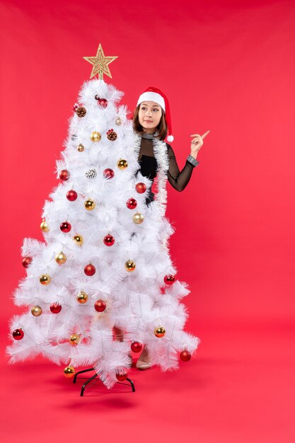 산타 클로스 모자가 크리스마스 트리 뒤에 숨어있는 검은 드레스에 아름다운 소녀와 함께 새해 분위기의 상위 뷰