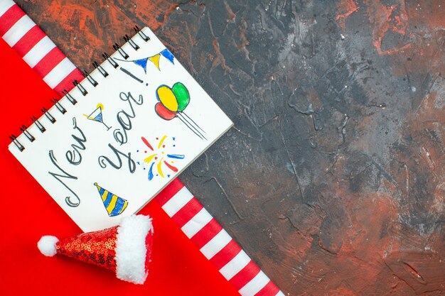 Top view new year written on notepad mini santa hat on red tablecloth on dark red table free space