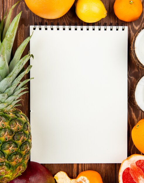 Top view of note pad with pineapple orange lemon tangerine coconut mango grapefruit around on wooden background with copy space