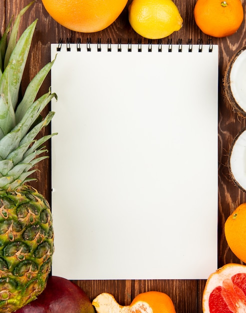 Top view of note pad with pineapple orange lemon tangerine coconut mango grapefruit around on wooden background with copy space