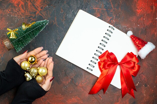 Top view notebook red bow xmas ball toys in female hands mini xmas tree small santa hat on dark red table