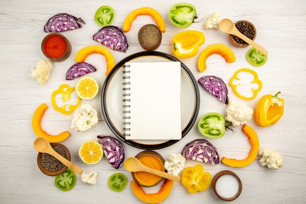 Top view notebook on round platter cut vegetables different spices in small bowls on white wooden surface