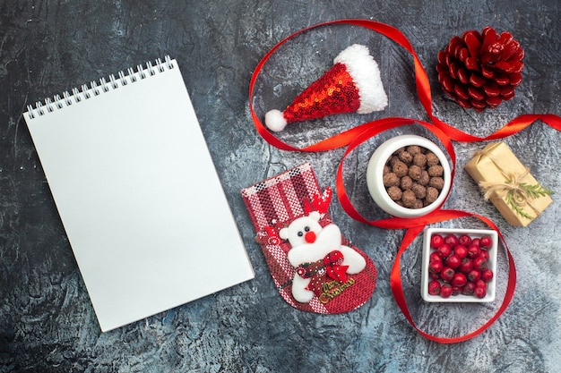 Free photo top view of notebook and santa claus hat and cornel chocolate new year sock red conifer cone on the left side on dark surface