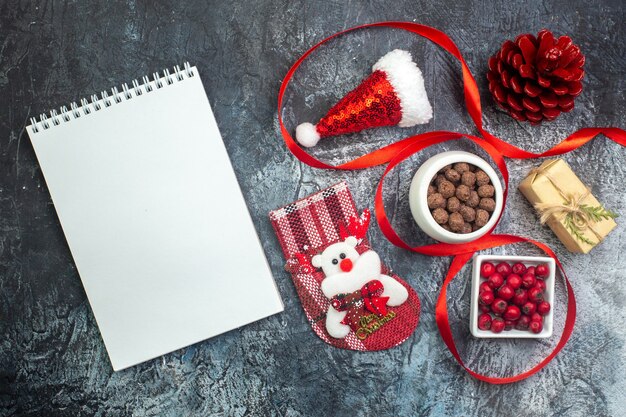 Top view of notebook and santa claus hat and cornel chocolate new year sock red conifer cone on the left side on dark surface