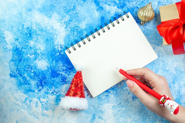 Top view notebook small santa hat red santa pen in female hand on blue white table