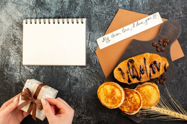 Top view of notebook and tasty breakfast with pancakes croisasant and hand opening gift box on dark table