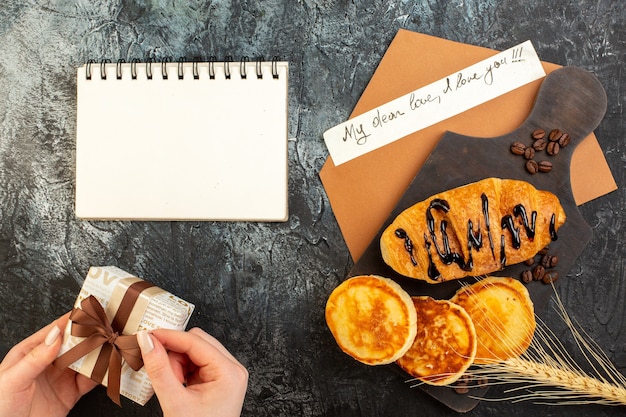 Top view of notebook and tasty breakfast with pancakes croisasant and hand opening gift box on dark table