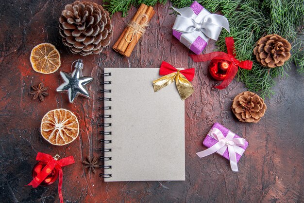 小さな弓の松の木の枝の円錐形のクリスマスツリーのおもちゃとギフトシナモンスティック乾燥レモンスライスと濃い赤の背景のノートブックの上面図
