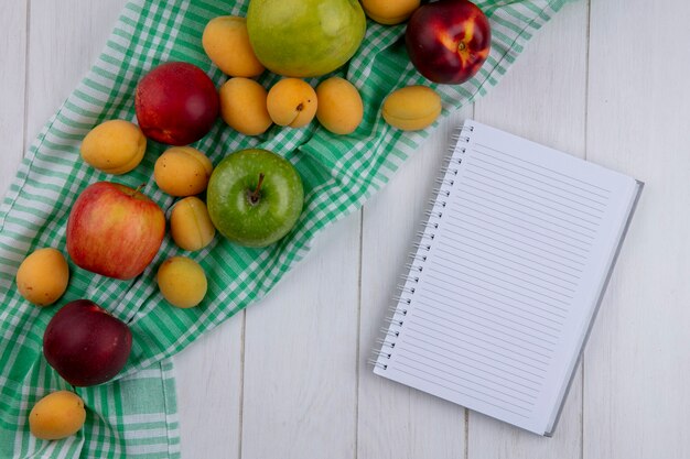 Вид сверху тетради с персиками, яблоками и абрикосами на клетчатом полотенце на белой поверхности