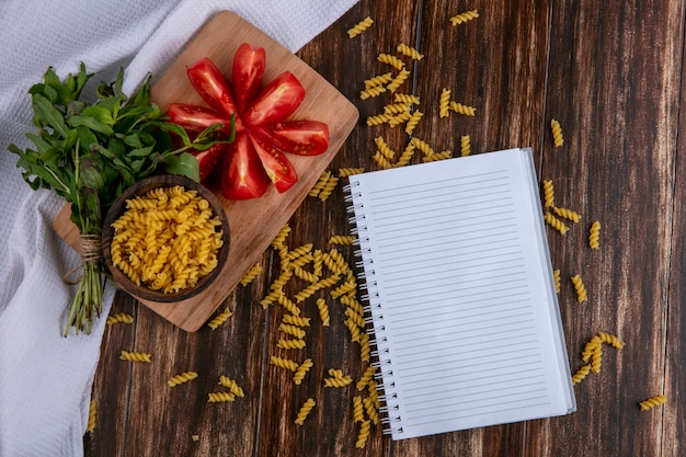 Top view of notebook with raw pasta in a bowl with slicing tomato slices on a cutting board with a bunch of mint on a wooden surface