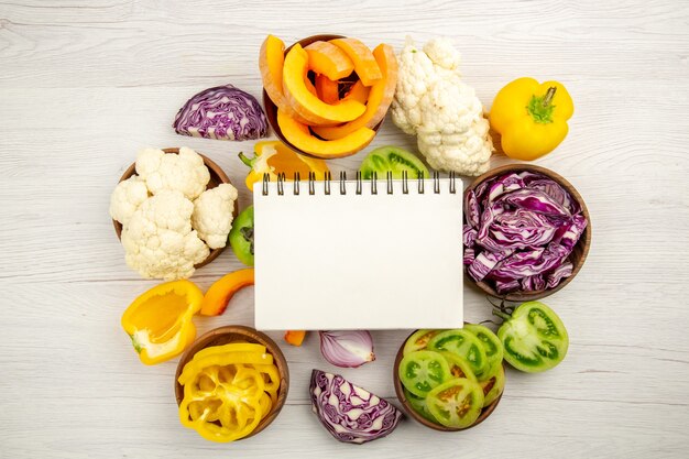 Top view notepad cut green tomatoes cut red cabbage cut pumpkin cauliflower cut bell peppers in bowls on surface