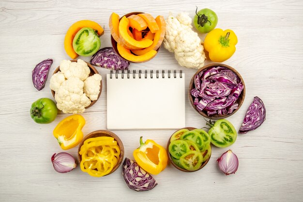 Top view notepad red cabbage cauliflower yellow bell pepper green tomato in bowls on white wooden surface