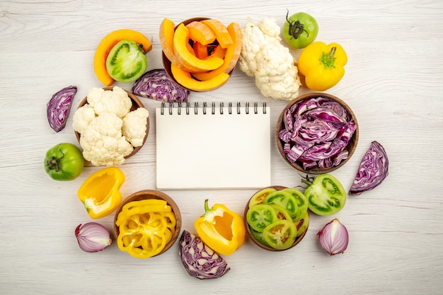 Top view notepad red cabbage cauliflower yellow bell pepper green tomato in bowls on white wooden surface