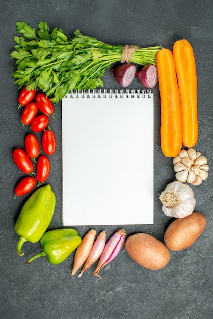 Top view of notepad with vegetables around it on dark grey background