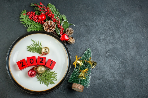 Vista dall'alto di accessori per la decorazione di numeri su un cono di conifera di rami di abete piatto accanto all'albero di natale su sfondo scuro