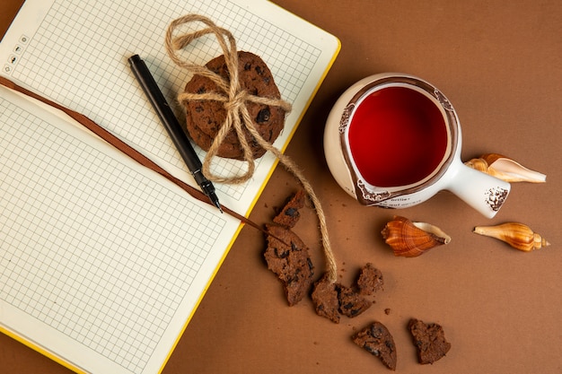 Top view of oatmeal cookies with chocolate chips and open empty notebook with pen and a cup of tea on ocher