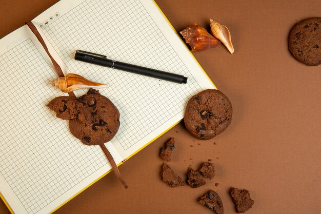 Top view of oatmeal cookies with chocolate chips and open empty notebook with pen on ocher