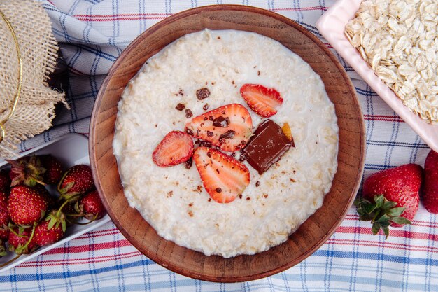 Top view of oatmeal porridge with fresh strawberries and chocolate in a wood bowl on plaid fabric