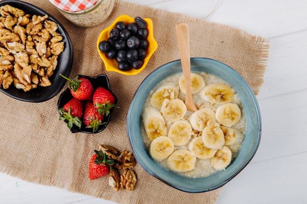 Top view of oatmeal porridge with sliced banana in a ceramic plate and fresh strawberries blueberry and nuts on sackcloth