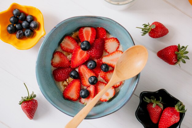 Top view of oatmeal porridge with sliced strawberries and blueberries in a ceramic bowl on the table