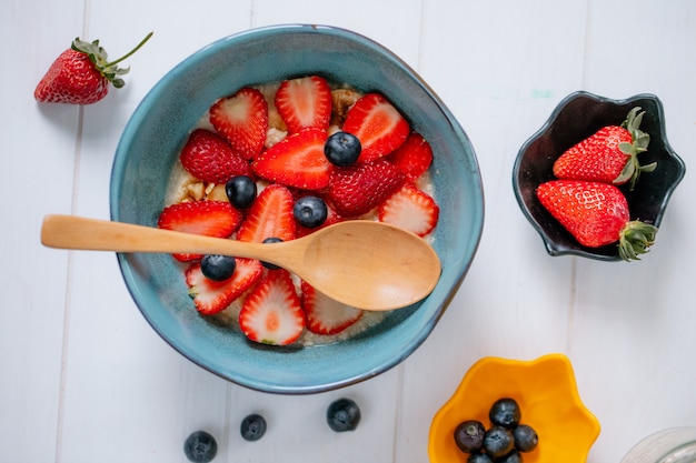 Top view of oatmeal porridge with sliced strawberries and blueberries in a ceramic bowl on white wooden surface