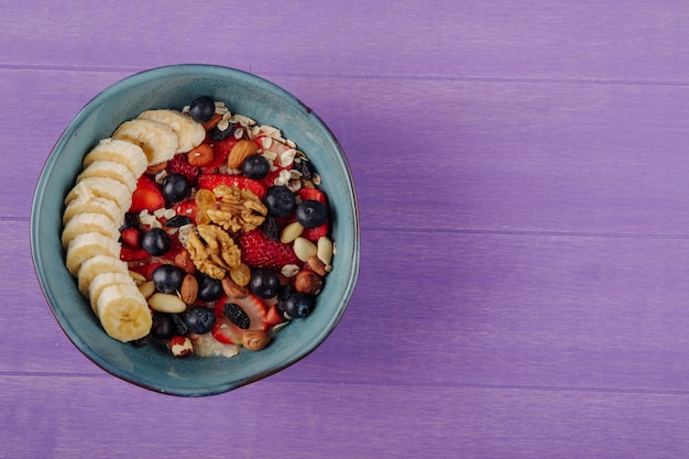 Top view of oatmeal porridge with strawberries blueberries bananas dried fruits and nuts in a ceramic bowl on purple wooden surface with copy space