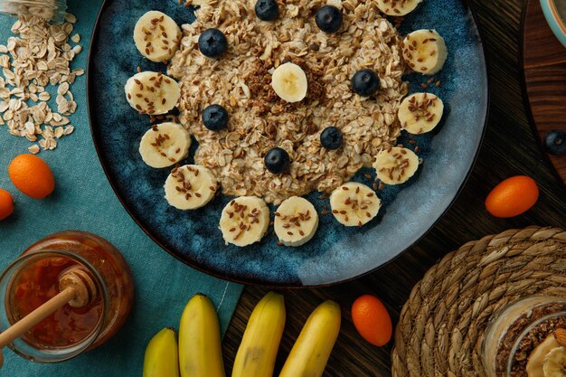 Top view of oatmeal with banana blackthorn walnut and sesame in plate with jam oat kumquat banana on blue cloth on wooden background