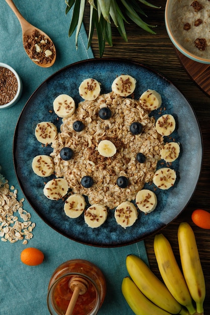 Free photo top view of oatmeal with banana blackthorn walnut and sesame in plate with jam oat kumquat pineapple leaves on blue cloth on wooden background