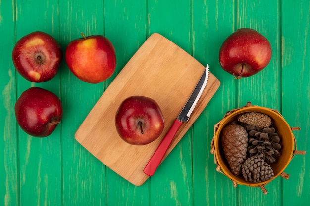 Бесплатное фото Вид сверху здорового красного яблока на деревянной кухонной доске с ножом с шишками на ведре с яблоками, изолированным на зеленой деревянной стене