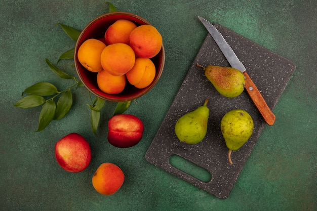Бесплатное фото Вид сверху абрикосов в миске с рисунком персиков и груш с ножом и листьями на разделочной доске и на зеленом фоне