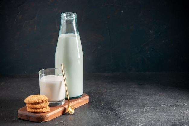 Вид сверху на вкусное молоко в стекле и бутылке с золотой ложкой, сложенной печеньем на деревянном подносе с правой стороны на темном фоне