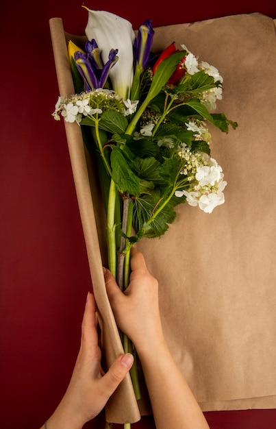 Бесплатное фото Взгляд сверху женских рук оборачивая букет цветущей калины лилии и темно-фиолетовых ирисов с крафт-бумагой на темно-красном столе
