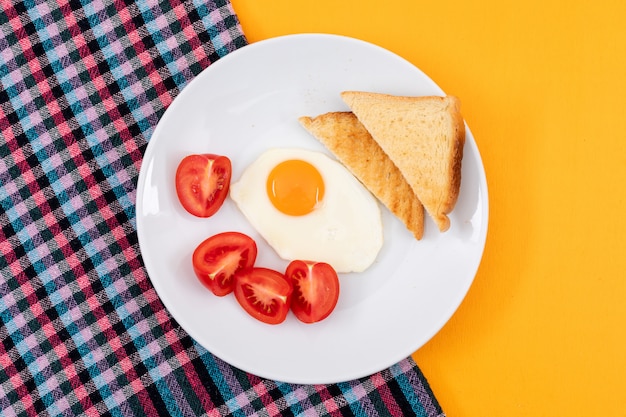 Бесплатное фото Вид сверху жареного яйца с тостами и помидорами на белой тарелке и желтой горизонтальной поверхности