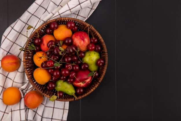 Вид сверху фруктов как вишнево-персик-абрикос-груша в корзине и на клетчатой ткани на черном фоне с копией пространства