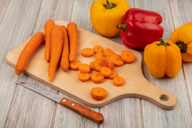 Вид сверху богатой витаминами и минералами овощной моркови на деревянной кухонной доске с ножом с разноцветным болгарским перцем, изолированным на серой деревянной поверхности