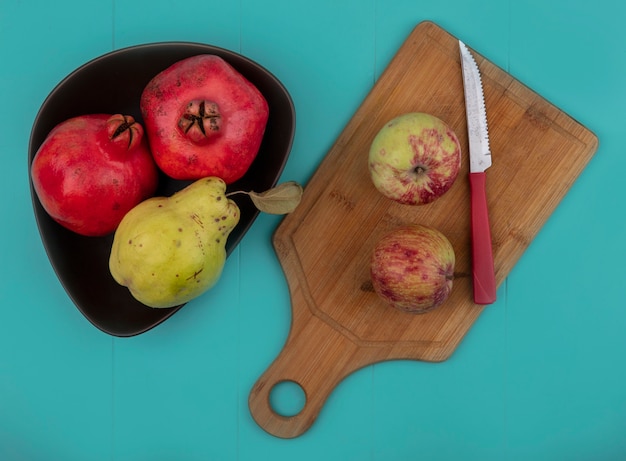 Бесплатное фото Вид сверху румяных свежих гранатов на миске с яблоками на деревянной кухонной доске с ножом на синем фоне
