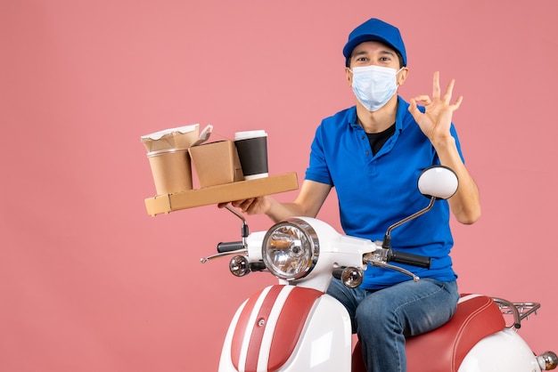 Вид сверху улыбающегося курьера в медицинской маске в шляпе, сидящего на скутере, делая жест очки на пастельном персике