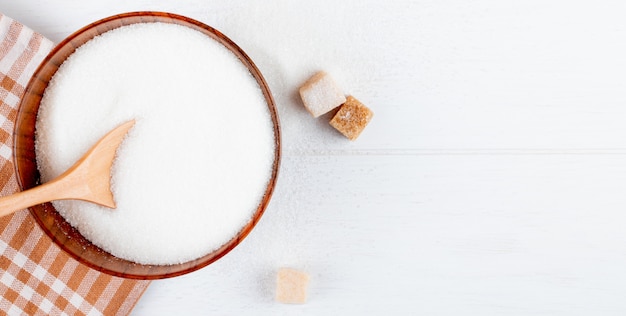 Бесплатное фото Вид сверху белого сахара в деревянной миске с ложкой и кусковой сахар на белом фоне с копией пространства