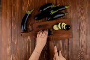 Бесплатное фото Взгляд сверху рук женщины режа баклажан с ножом на разделочной доске с целыми на деревянной предпосылке