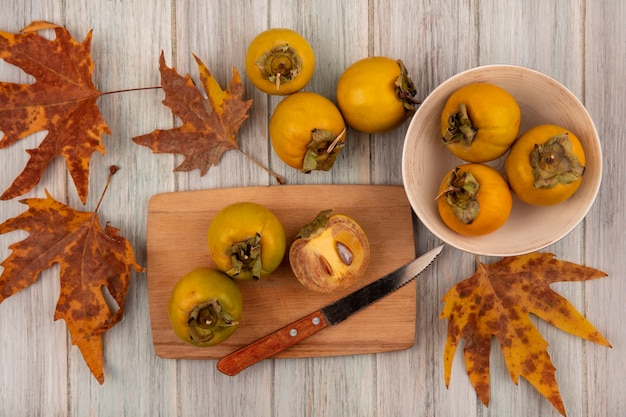 Бесплатное фото Вид сверху желтых плодов хурмы на миске с листьями с фруктами хурмы на деревянной кухонной доске с ножом на сером деревянном столе