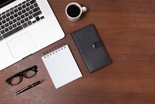 Top view of office desk with hot tea; blank spiral notepad; diary; eyeglasses; pen and laptop