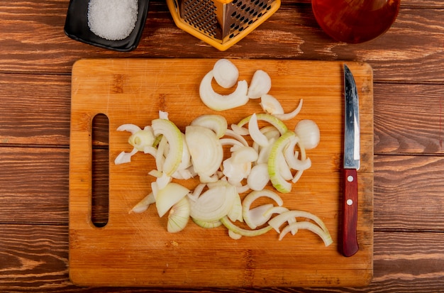 Top view of onion slices and knife on cutting board with salt butter and grater on wooden background