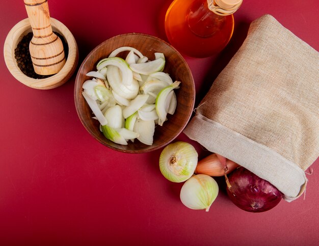 Top view of onions spilling out of sack with sliced ones in bowl and melted butter with black pepper seeds in garlic crusher on red