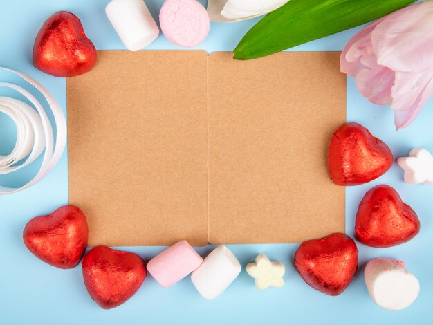 Top view of an open brown paper greeting card with scattered marshmallows and heart shaped chocolate candies in red foil with pink color tulips on blue table