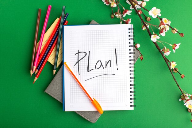 Top view open copybook with colorful pencils and flowers on green surface