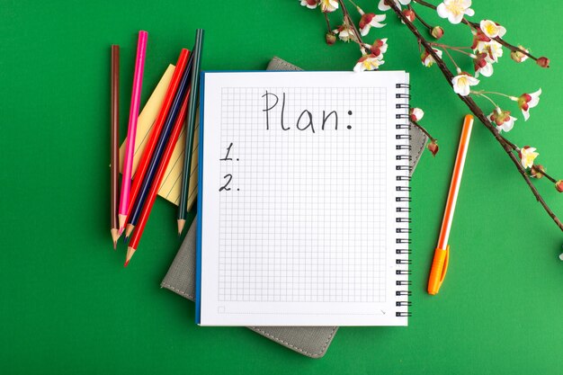 Top view open copybook with colorful pencils and flowers on the green surface